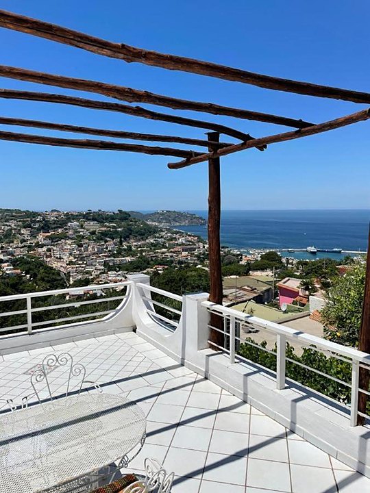 Terrazza bianca sotto un pergolato rustico, con Casamicciola e il mare